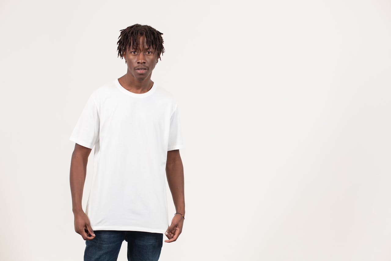 Serious young African American male model in simple white t shirt and jeans standing in light studio and looking at camera